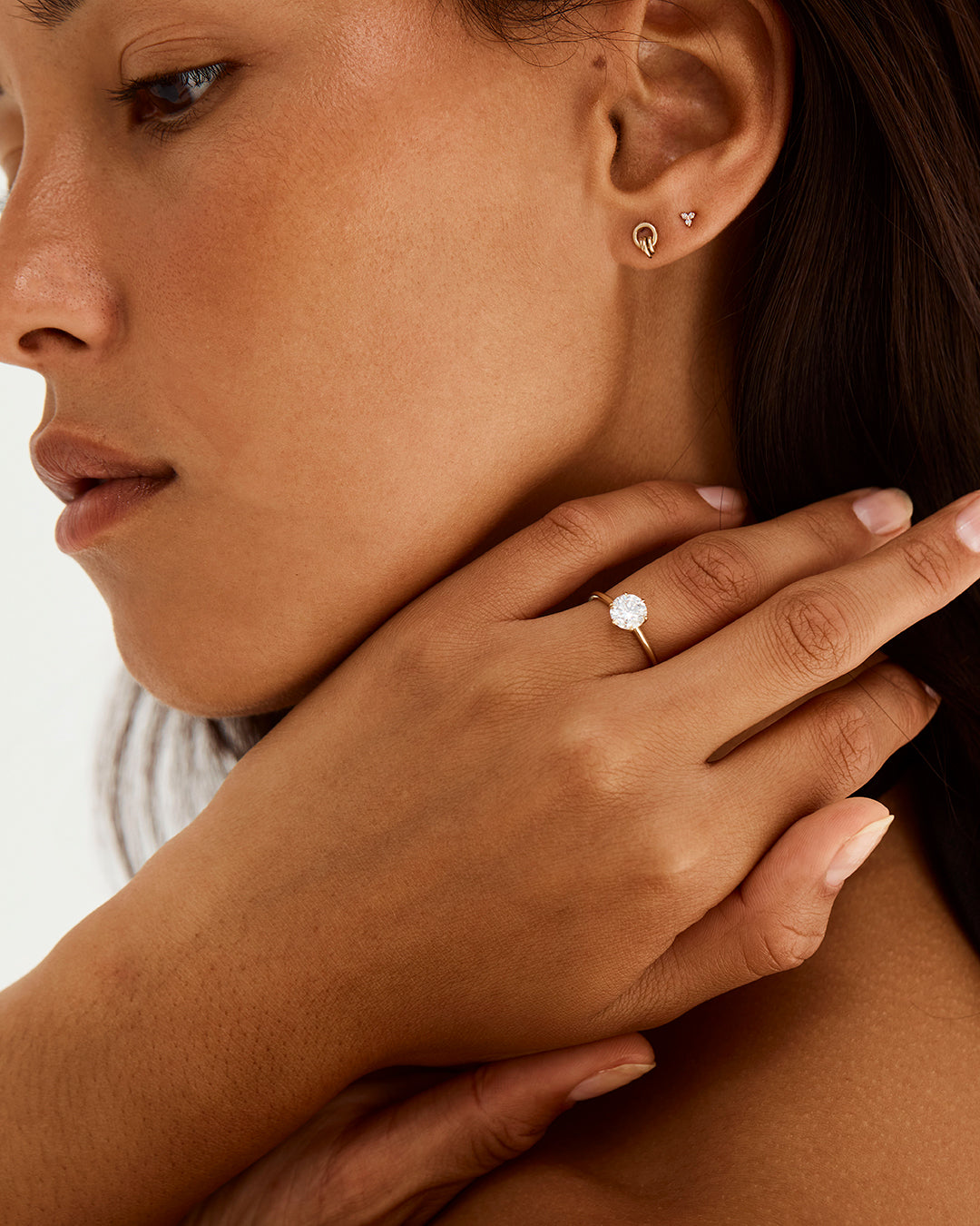 A model wears a solitaire style engagement ring, featuring a round cut white diamond, with diamonds in and around the setting