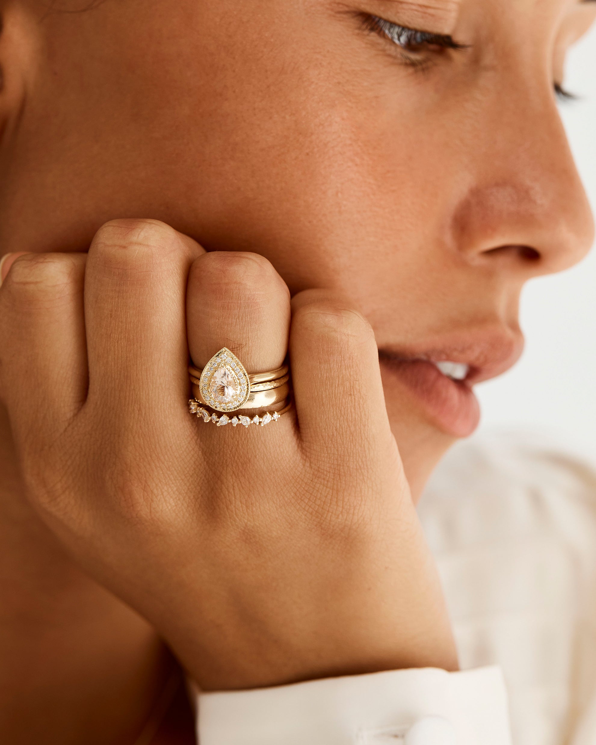 Model wears a pear halo engagement ring stacked with our fine faceted texture band, classic gold band and pear and round alternating diamond ring.