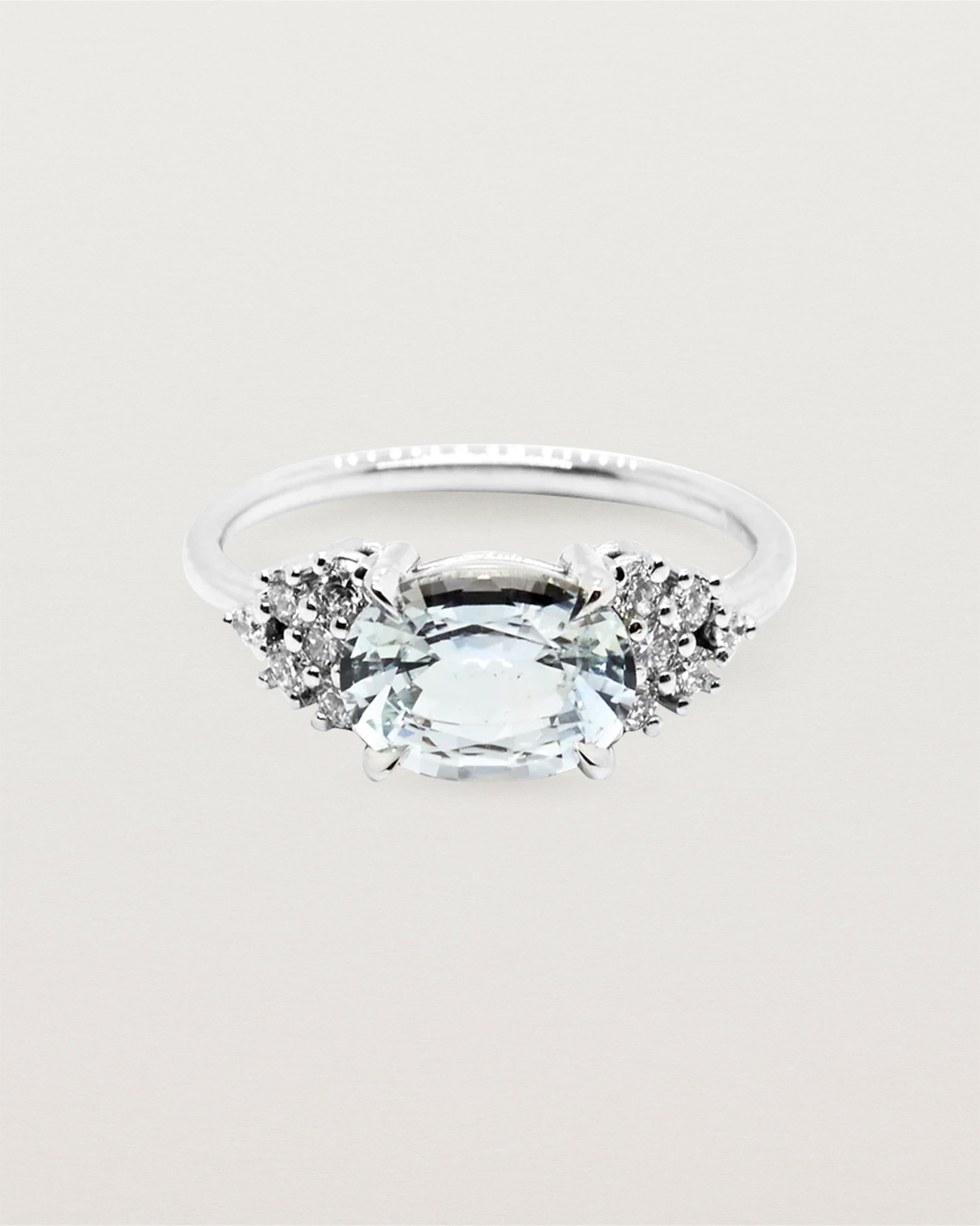 A white oval sapphire set east to west, adorned with clusters of white diamonds either side, and crafted in white gold