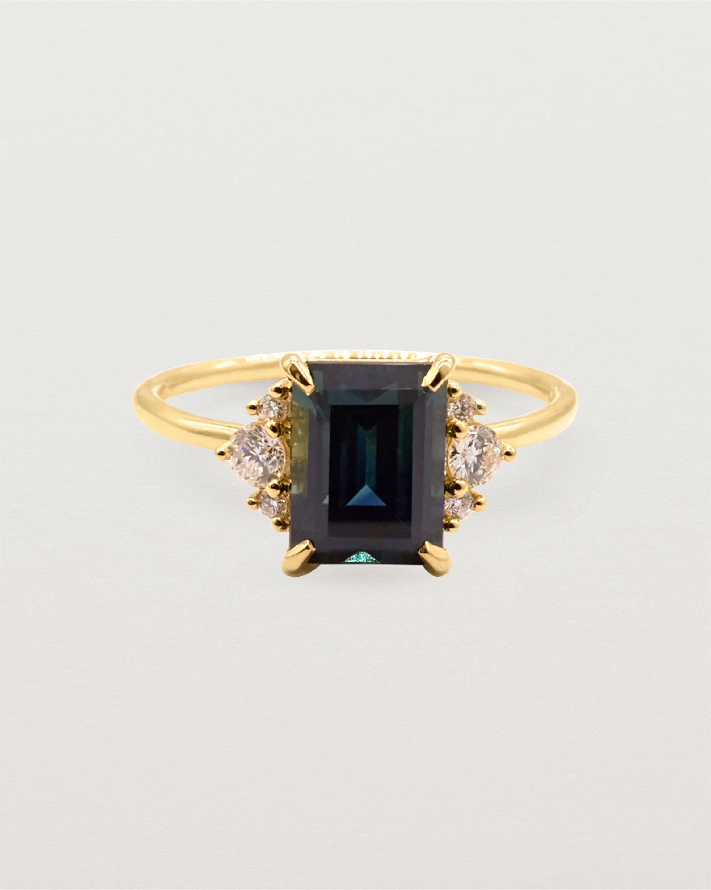 A stunning blue/green emerald cut parti sapphire set amongst a delicate trio cluster of round white diamonds. Featuring a round band and intricate basket setting all in 18ct Yellow Gold.