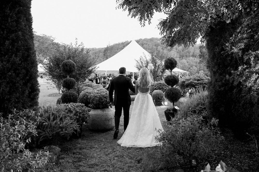 A groom and bride walking along a walkway on grass hand in hand with greenery on each side