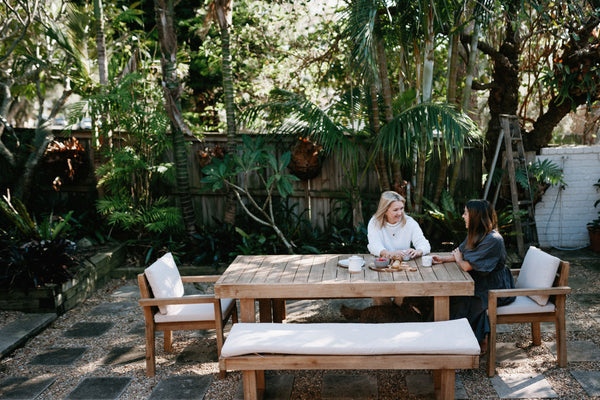 A women in a white jumper sitting at a big wooden outdoor table in a lush backyard