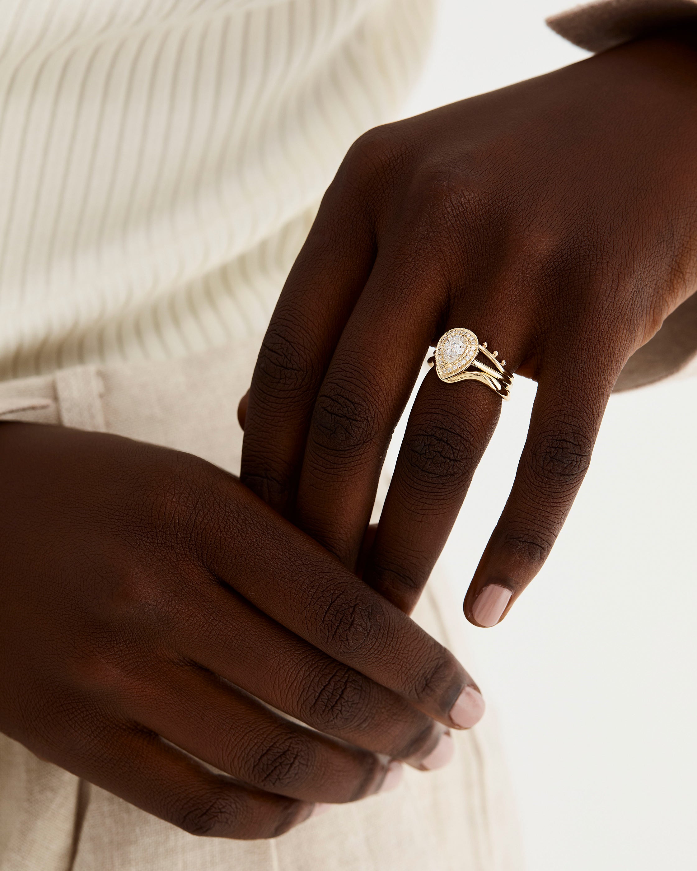 A model wears a pear cut white diamond halo engagement ring stacked with diamond and plain wedding bands