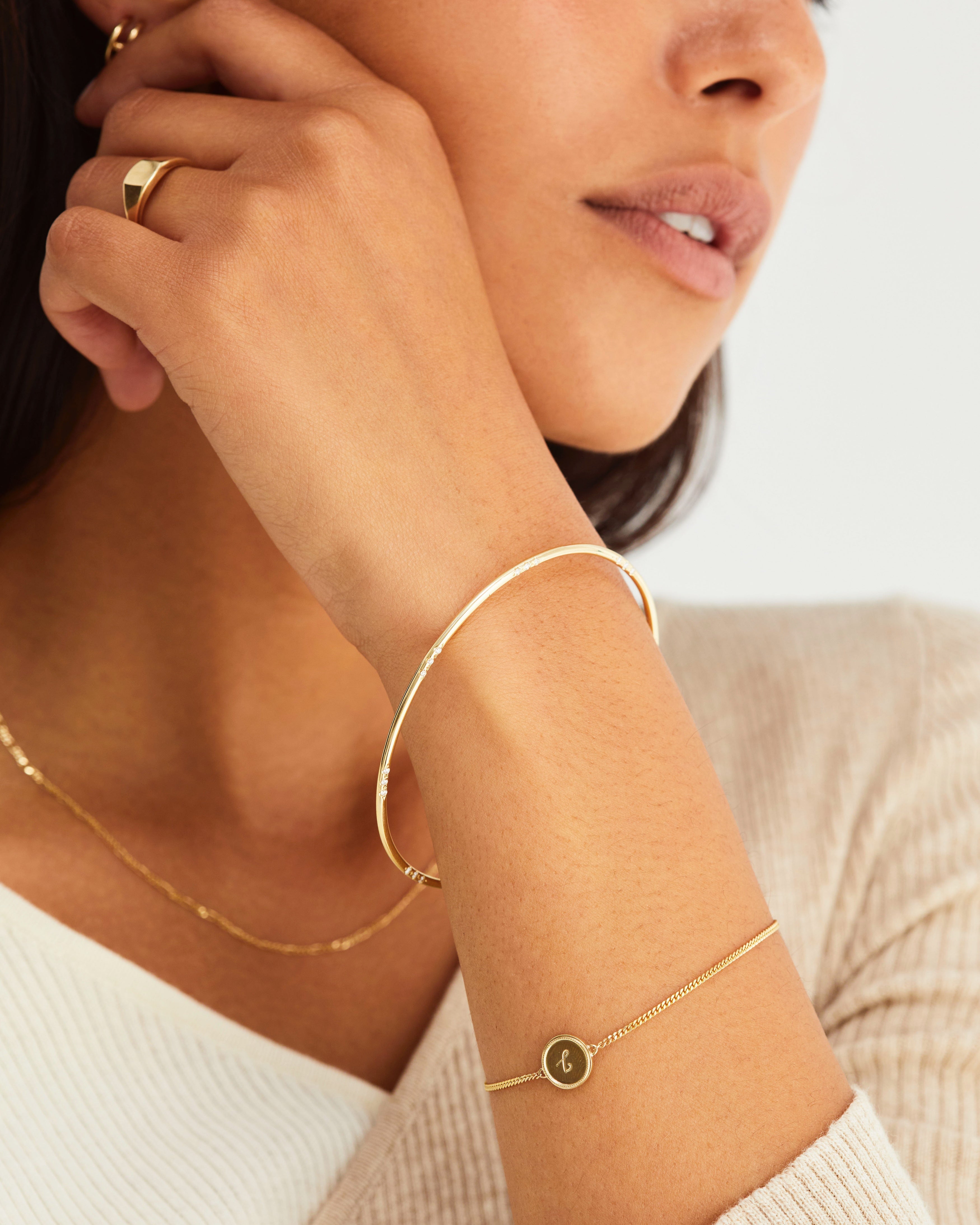A woman wearing the Precious Initial Bracelet | Birthstone and the Cascade Oval Bangle | Diamonds, both in yellow gold.