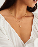 A model wears a heart shaped pendant necklace layered with a small knot necklace in yellow gold
