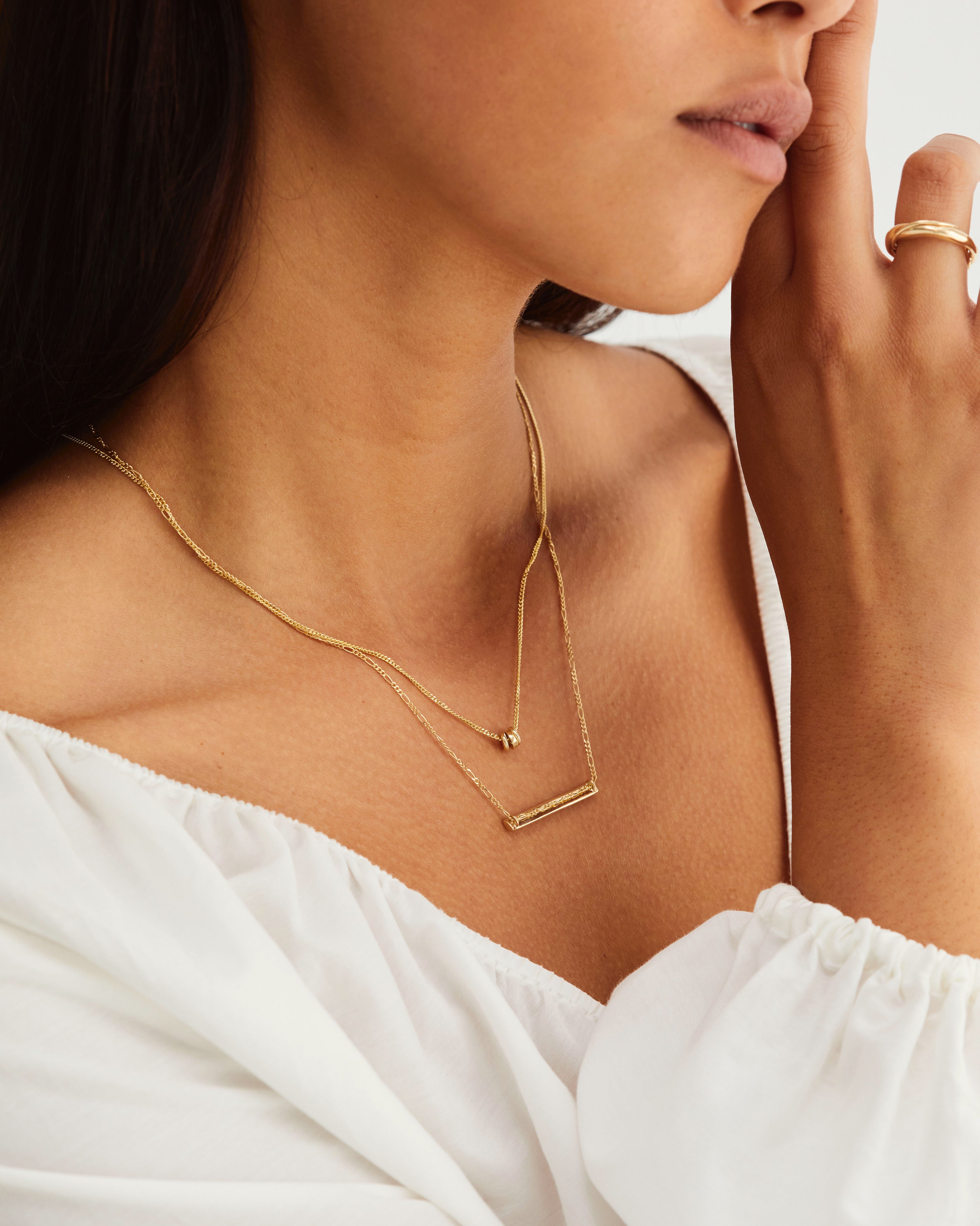 A model wears a bar shaped necklace on a detailed chain, layered with a small golden charm necklace