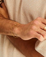 A man wears the Aether Bracelet layered with a Figaro chain, in yellow gold