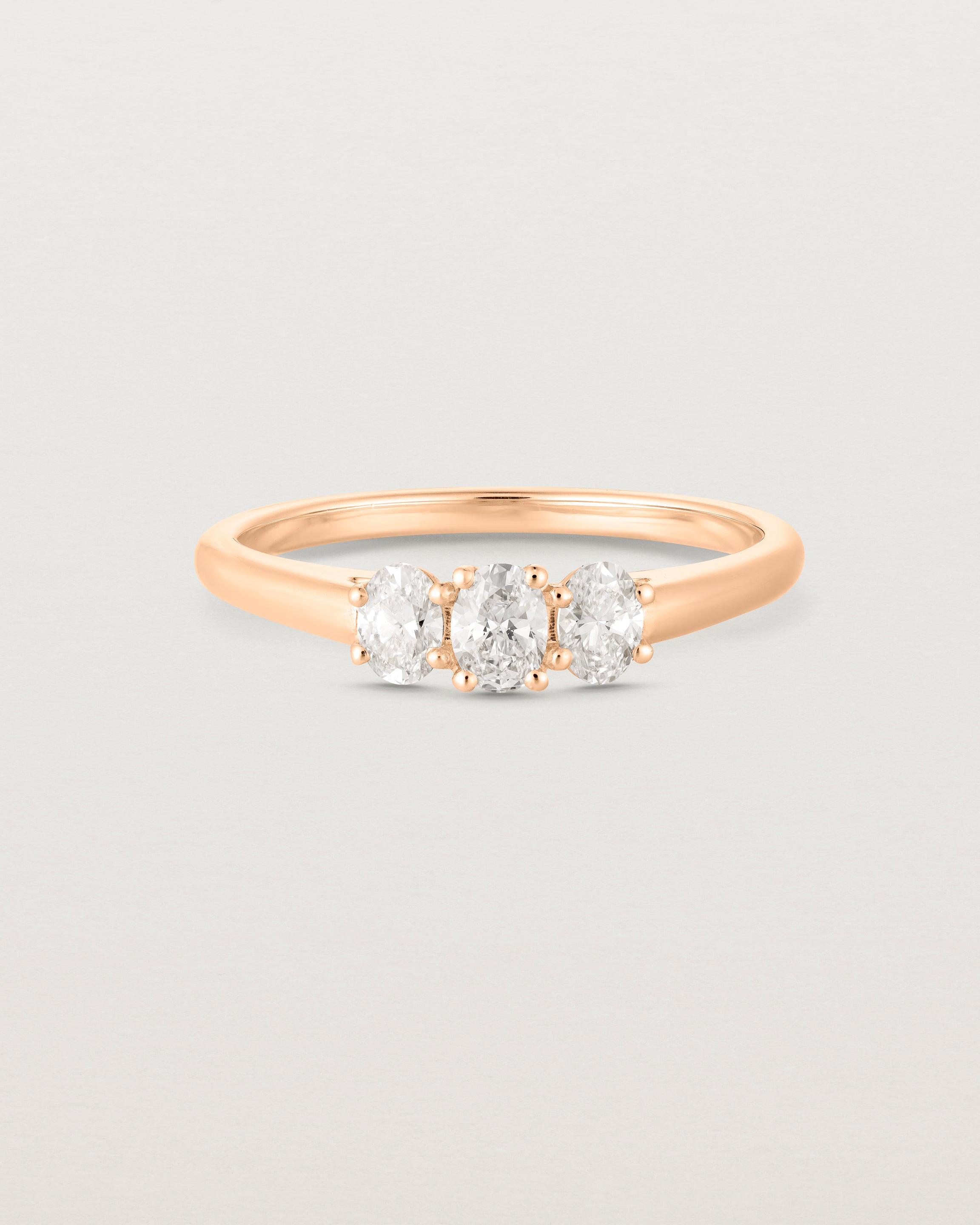 Front on deep etched image of a rose gold engagement ring with three oval white laboratory grown diamonds.