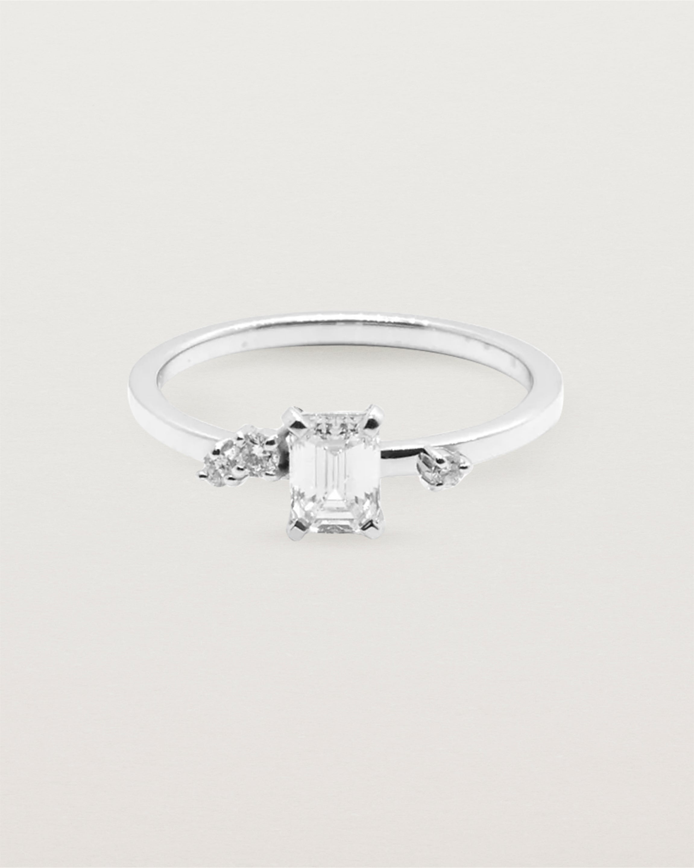A petite emerald cut white diamond sits atop a square band with white diamond clusters either side, crafted in white gold.