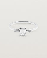 A petite emerald cut white diamond sits atop a square band with white diamond clusters either side, crafted in white gold.