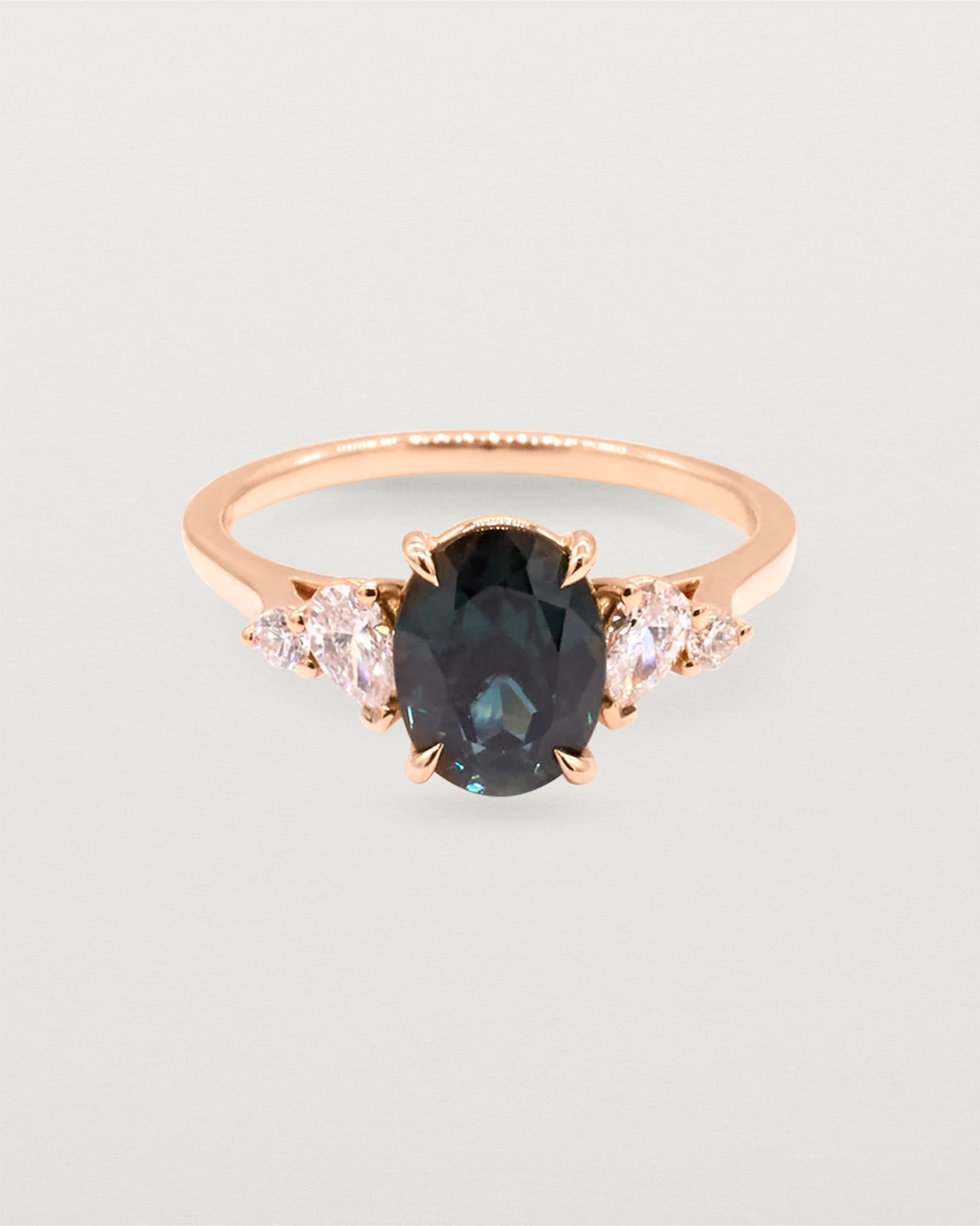 A deep Australian teal sapphire sits within a four claw setting, framed by a delicate cluster of pear and round white diamonds atop a rose gold soft square band.
