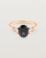 A deep Australian teal sapphire sits within a four claw setting, framed by a delicate cluster of pear and round white diamonds atop a rose gold soft square band.