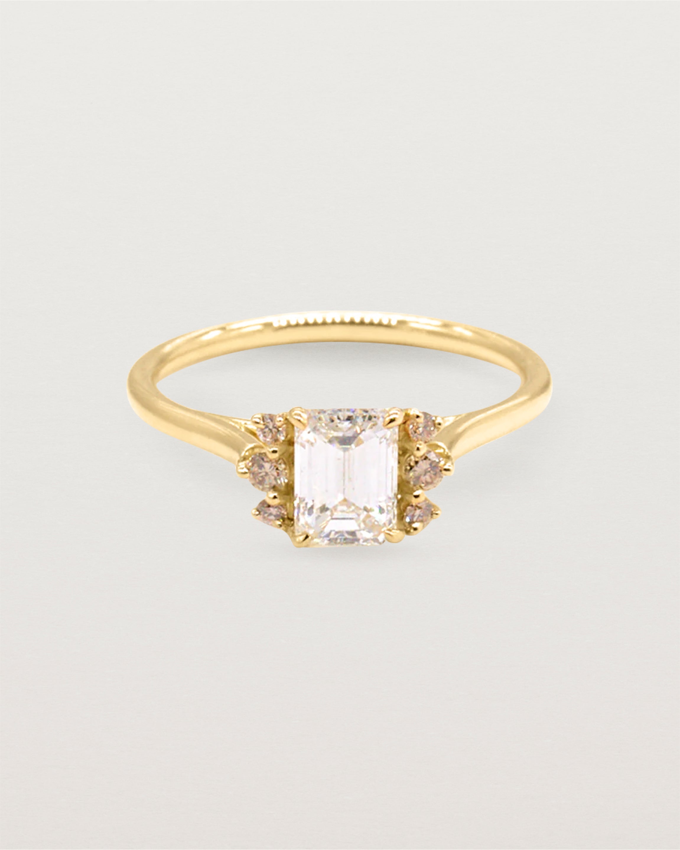 A emerald cut white diamond is shouldered by sparkling champagne diamonds and crafted in yellow gold. 