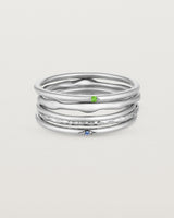 A set of five white gold bands, one featuring a blue sapphire, one featuring a green emerald, one textured and two plain
