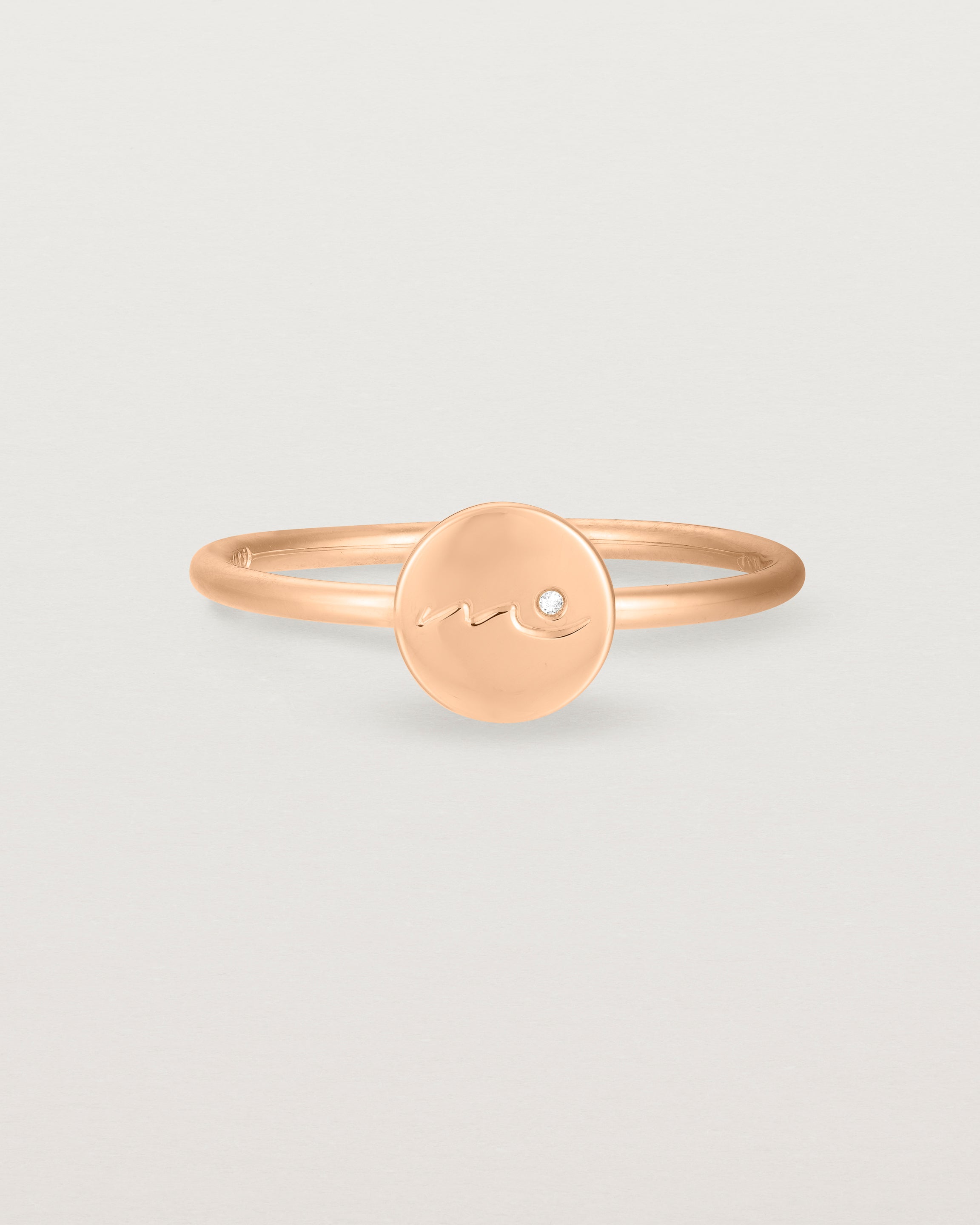 Front view of a rose gold ring featuring a disc with the letter m engraved and a small diamond set